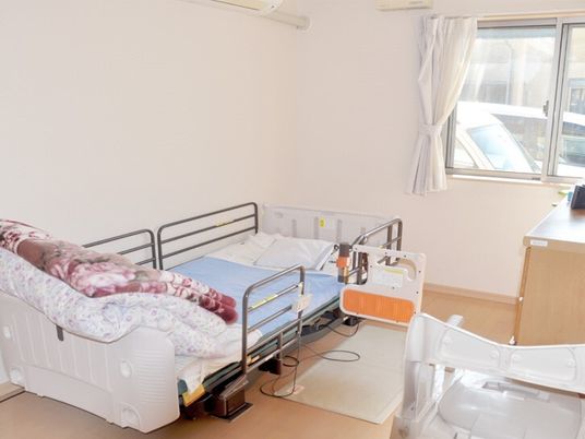 寝具付きのベッドが置かれたフローリングの居室にはタンスやポータブルトイレもあり、大きな窓が付き明るい雰囲気。