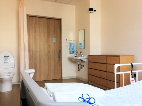 フローリングの居室にはカーテンで開け閉めをするタイプのトイレが設置されている。ベッドが置かれ、収納家具も設置されている。