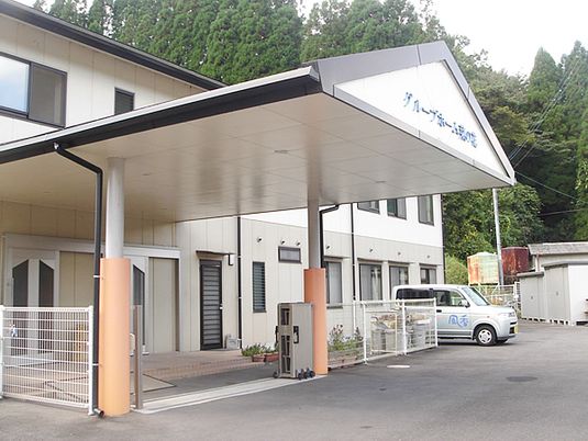 大きな車寄せが付いた白を基調とした2階建ての建物。駐車スペースには事業所名が書かれた車が止められている。