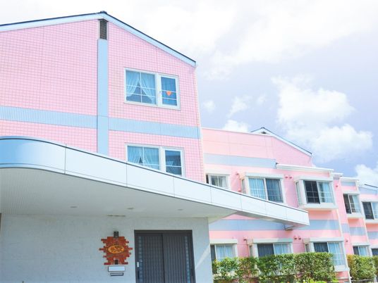 ３階建ての建物はピンクで周辺には緑が植えられている。