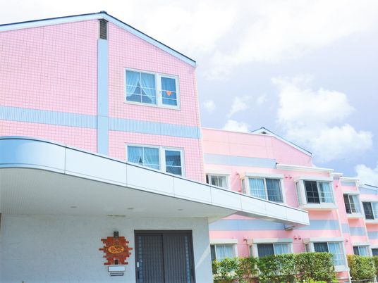 ３階建ての建物はピンクで周辺には緑が植えられている。