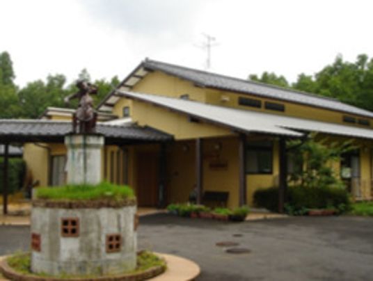 ベージュの外壁の平屋建ての建物にはグレーの瓦屋根が付き、玄関先にはベンチが置かれている。