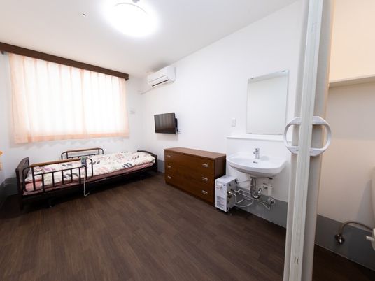 トイレ付きのフローリングの部屋にはベッドや洗面所、テレビ、タンスなどがあり、大きな窓も付いている。