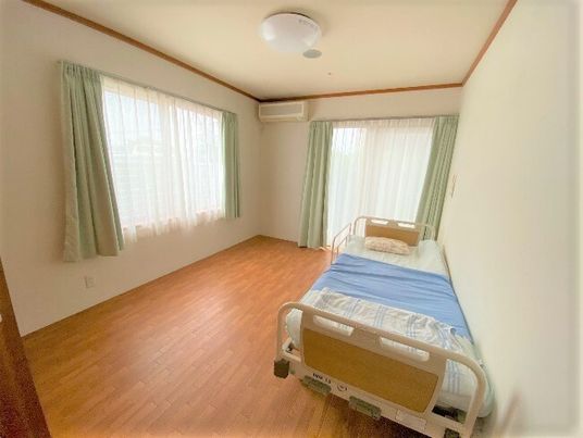 寝具付きのベッドが壁際に置かれた居室。大きな窓が二つあり、エアコンも壁についている。