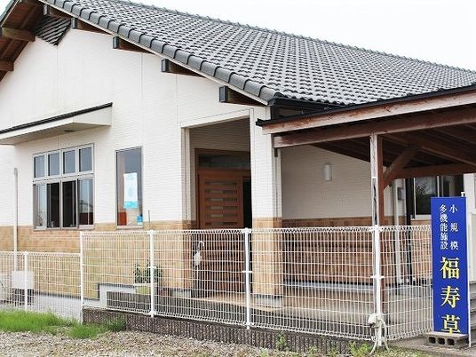 瓦屋根が付いた和風の建物。玄関の横には花が植えられたプランターが置かれ、フェンスにはホーム名が書かれた看板がある。