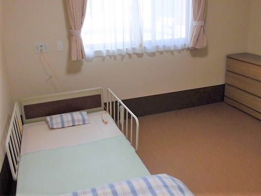 寝具がセットされた介護ベッドが置かれた居室には緊急通報装置が設置され、木製のタンスやカーテンが付いた大きな窓もある。