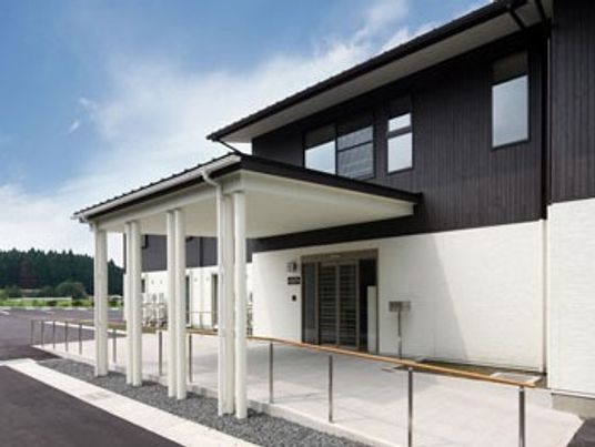 2階建ての建物はモノトーンでまとめられ、玄関付近には車寄せと手すりが付き、周辺を自然に囲まれている。