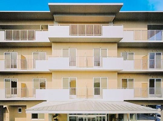 ベージュ系の4階建ての建物の各フロアにベランダがあり、1階部分と4階部分には屋根が付いている。