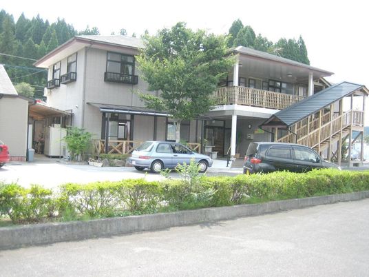 屋外階段を設置した2階建ての建物がある敷地内には駐車スペースがあり車が数台止められている。周りには緑が多く、のどかな環境。