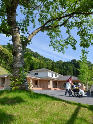 山々に囲まれた施設を外観から写したもの。車いすを押す利用者とスタッフの様子