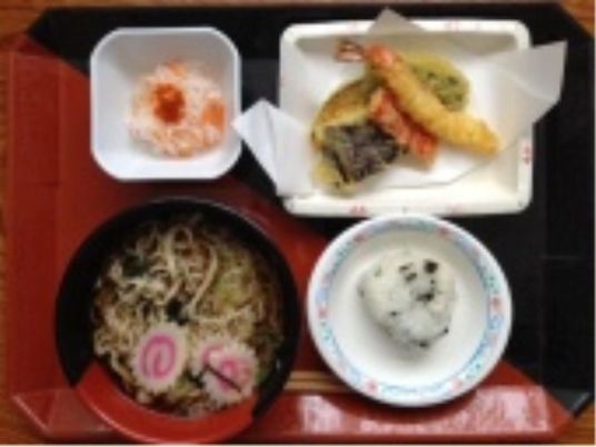 おにぎりにそば、天ぷらなどバランスのとれた食事が赤と黒のお盆に盛られている。