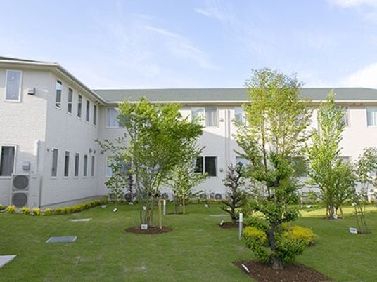 白い建物は2階建てでエル字型。広い庭には背の高い木がたくさん植えられていて、芝生も敷かれている。