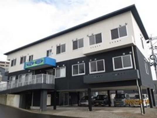 白と黒の3階建ての建物の1階部分は駐車場となっていて、玄関にはホーム名が書かれた屋根がある。