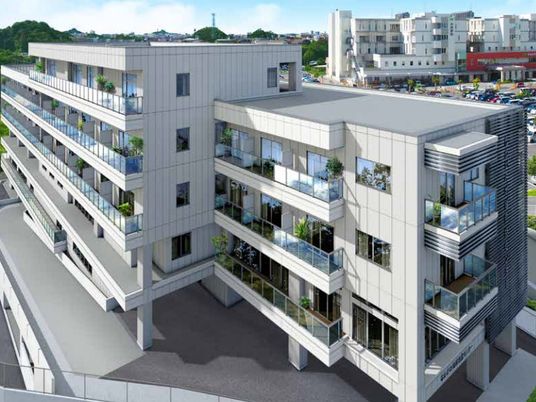 白を基調とした大きな建物の各フロアにベランダがあり、周囲には緑も多い。奥にも白い大きな建物がある。