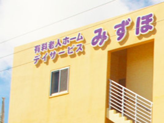 山吹色の外壁の建物にはホーム名と併設事業所名が書かれている。屋外階段が付いている。