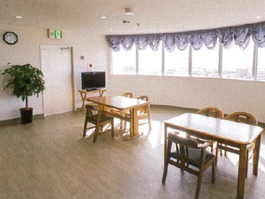 木製の3人掛けのテーブルが二つ置かれ、観葉植物やテレビ、時計などが飾られている食堂。