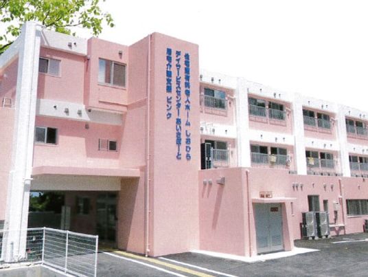 3階建ての建物は全体がピンク、白のラインが入り、館内の事業所の名前が青で書かれている。