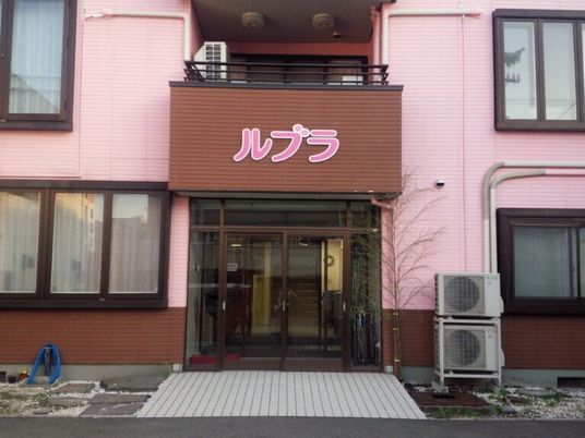 ブラウンとピンクの２階建ての建物玄関には自動ドアが付き、2階のバルコニー部分が玄関の屋根となっている。