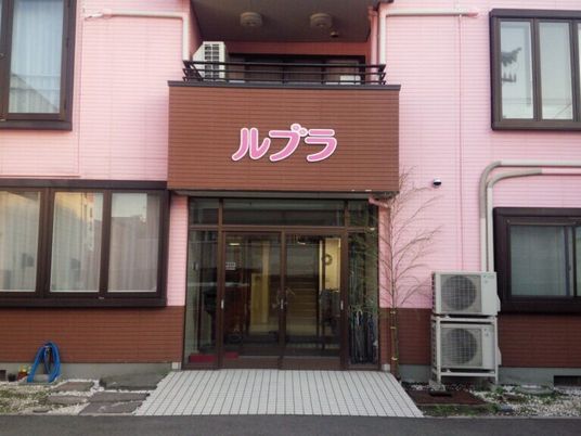 ブラウンとピンクの２階建ての建物玄関には自動ドアが付き、2階のバルコニー部分が玄関の屋根となっている。