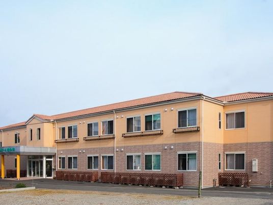 2階建ての建物はベージュ系で明るい雰囲気。横に長く、中央の玄関には車寄せが付けられている。