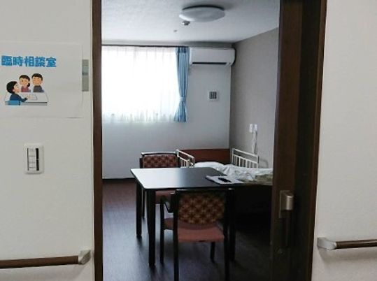 机と椅子が置かれた居室の入り口。臨時相談室の紙が貼ってあるスペース