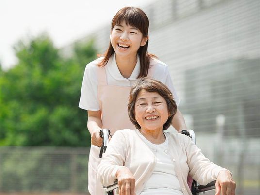 カーディガンを着た高齢女性と屋外で笑顔で過ごしているエプロンを付けた女性スタッフ。高齢女性は車いすに座り微笑んでいる。
