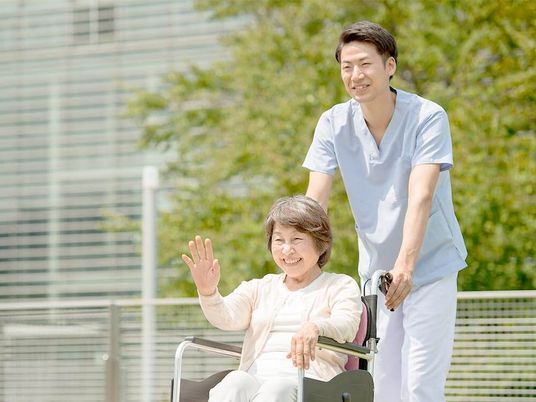 男性スタッフに車いすを押してもらいながら屋外を散歩している高齢女性。誰かに向かって手を振っている。