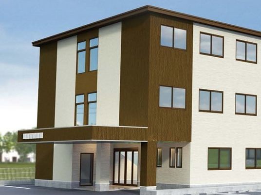 白とブラウンの３階建ての建物。車寄せが付き、周辺には緑が多い。