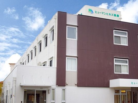 白とブラウンの３階建ての建物があり、緑の文字で施設名が外壁に書かれている。