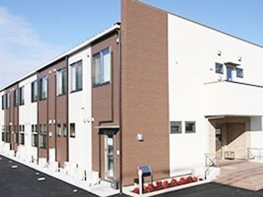 ブラウンと白の２階建ての建物は日当たりのいい場所に建っている。玄関には手すり付きの階段とスロープがある。