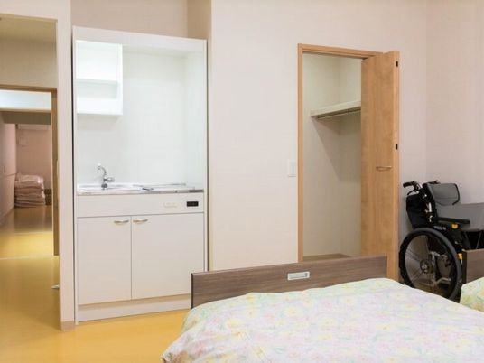 収納やキッチンがついた施設内の居室の様子。車いすとベッドが手前に置いてある様子