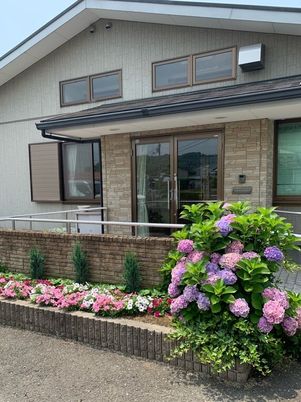 一般の住宅と変わらない雰囲気の三角屋根の平屋建て。周辺にはたくさんの花が咲き、明るい雰囲気。