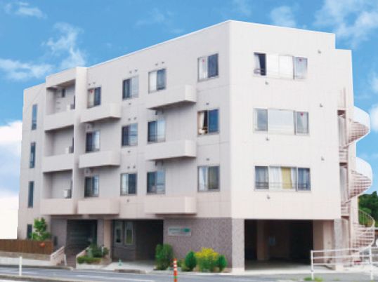 白を基調とした4階建ての建物は日当たりのいい場所に建っていて、屋外階段が付き、建物の周りには草木が植えられている。