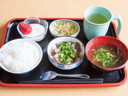 白いご飯にメインの肉料理、みそ汁やデザートまで付いた食事。黒いトレーには箸とスプーンも添えられている。