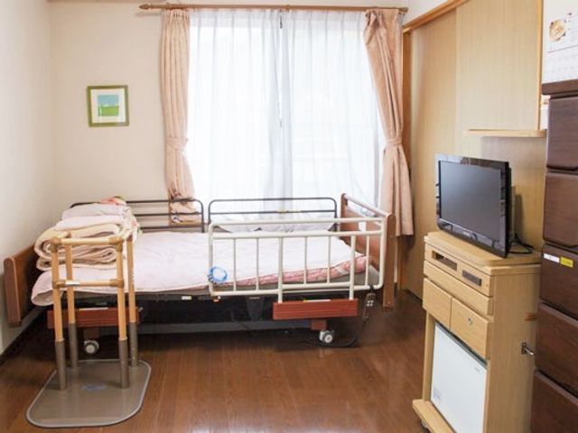 寝具付きのベッドが置かれたフローリングの居室には家具もあり、大きな窓が付き明るい雰囲気。 在宅での生活のお手伝いからお任せ！長期にわたって信頼関係を築いて行けるよう努めております。