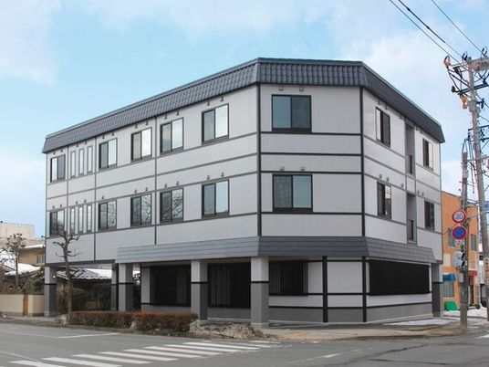 グレー系の3階建ての建物の周りには草木が植えられている。道路に面した場所に建っている。