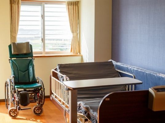 介護ベッドや車いすが置かれているフローリングの部屋にはカーテンが付いた大きな窓がある。