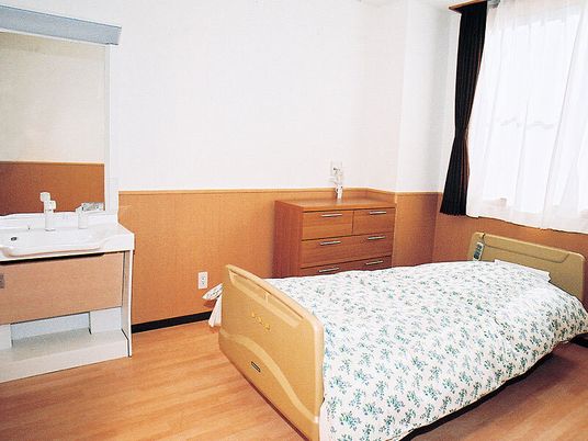 車いす対応の洗面所や介護用ベッド、タンスが置かれた部屋にはレースカーテンとカーテンが付いた窓もある。