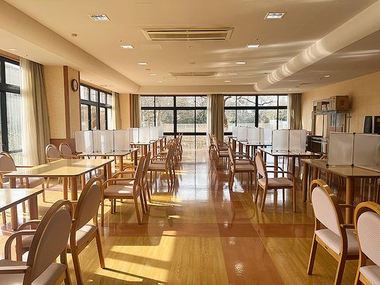 パーテーションが設置された木製のダイニングテーブルが並んだ食堂。大きな窓がたくさんついている。