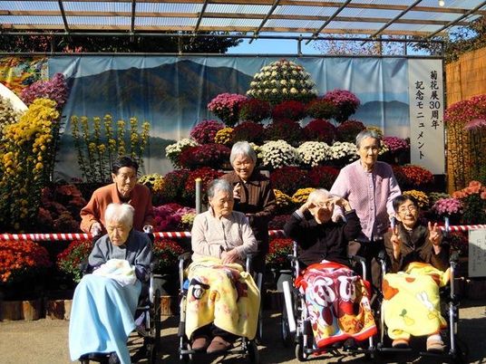 菊の花でできたオブジェの前で写真に納まる高齢者。車いすに乗っている人が手前に写っている様子