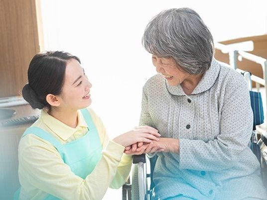 エプロンを付けた若い女性スタッフと車いすの高齢女性が笑顔で手を重ねて話している。