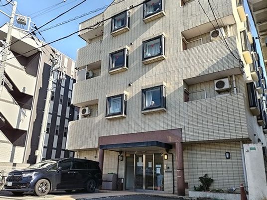 車が止まっている駐車場がある4階建てのベージュの外壁の建物。日当たりのいい場所に建っている。