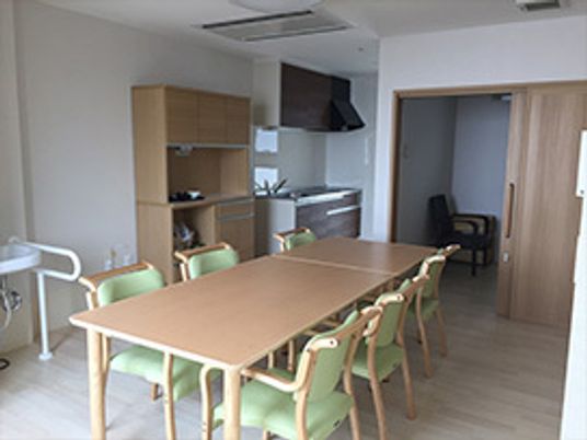 ライトグリーンの椅子がセットされた木製のテーブルや車いす対応の洗面所、キッチンがあるスペース。