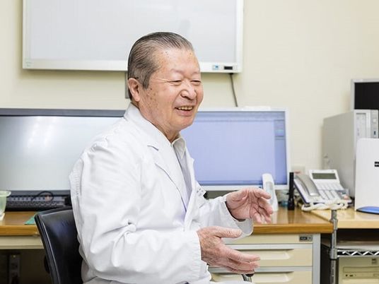 白衣を着た高齢の男性医師が笑顔で話している。後ろのデスクにはパソコンや電話がある。