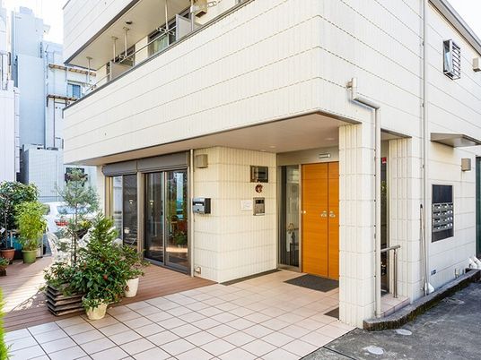 白い外壁の建物の玄関館にはインターホンがあり、マットが敷かれ、草木が植えられた鉢がたくさん置かれている。