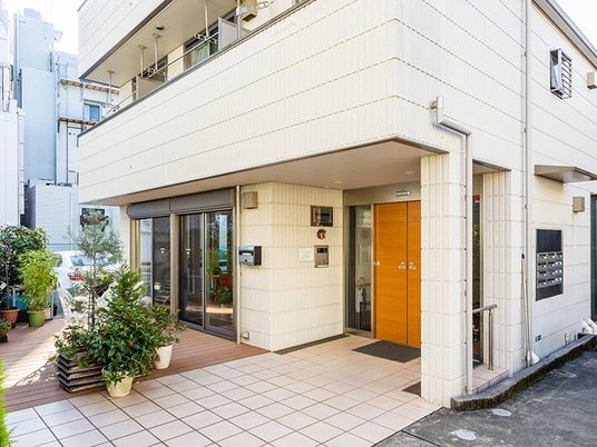 白い外壁の建物の玄関館にはインターホンがあり、マットが敷かれ、草木が植えられた鉢がたくさん置かれている。