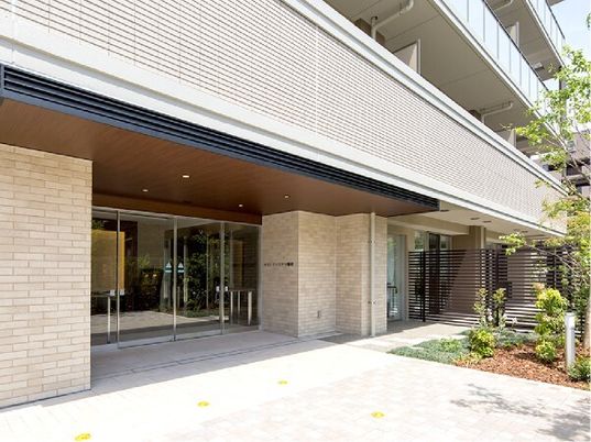 広い駐車場が付いた2階建ての建物。建物の周辺には緑が多くあり、玄関には車寄せも付いている。