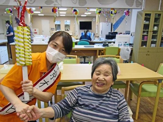 「東京オリンピック」と書かれたたすきをかけて高齢女性と手を握り合っているポロシャツ姿の女性スタッフ。