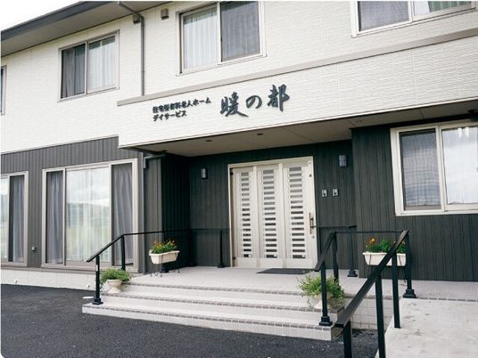 モノトーンの2階建ての建物の玄関には階段とスロープが付き、ホーム名と併設のデイサービスの名前が書かれている屋根も設置されている。