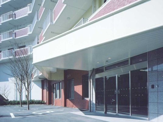 白とピンクの外観の建物の各フロアにベランダがあり、玄関には車寄せがあり、駐車場も付いている。